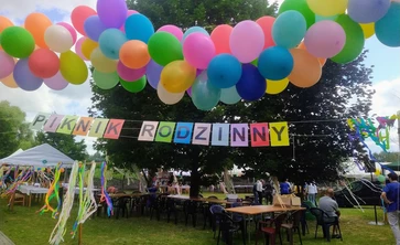 zdjęcie ogrodu udekorowany napisem piknik rodzinny, kolorowe balony, drzewa w tle stoły krzesła