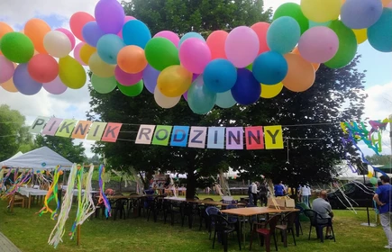 zdjęcie ogrodu udekorowany napisem piknik rodzinny, kolorowe balony, drzewa w tle stoły krzesła